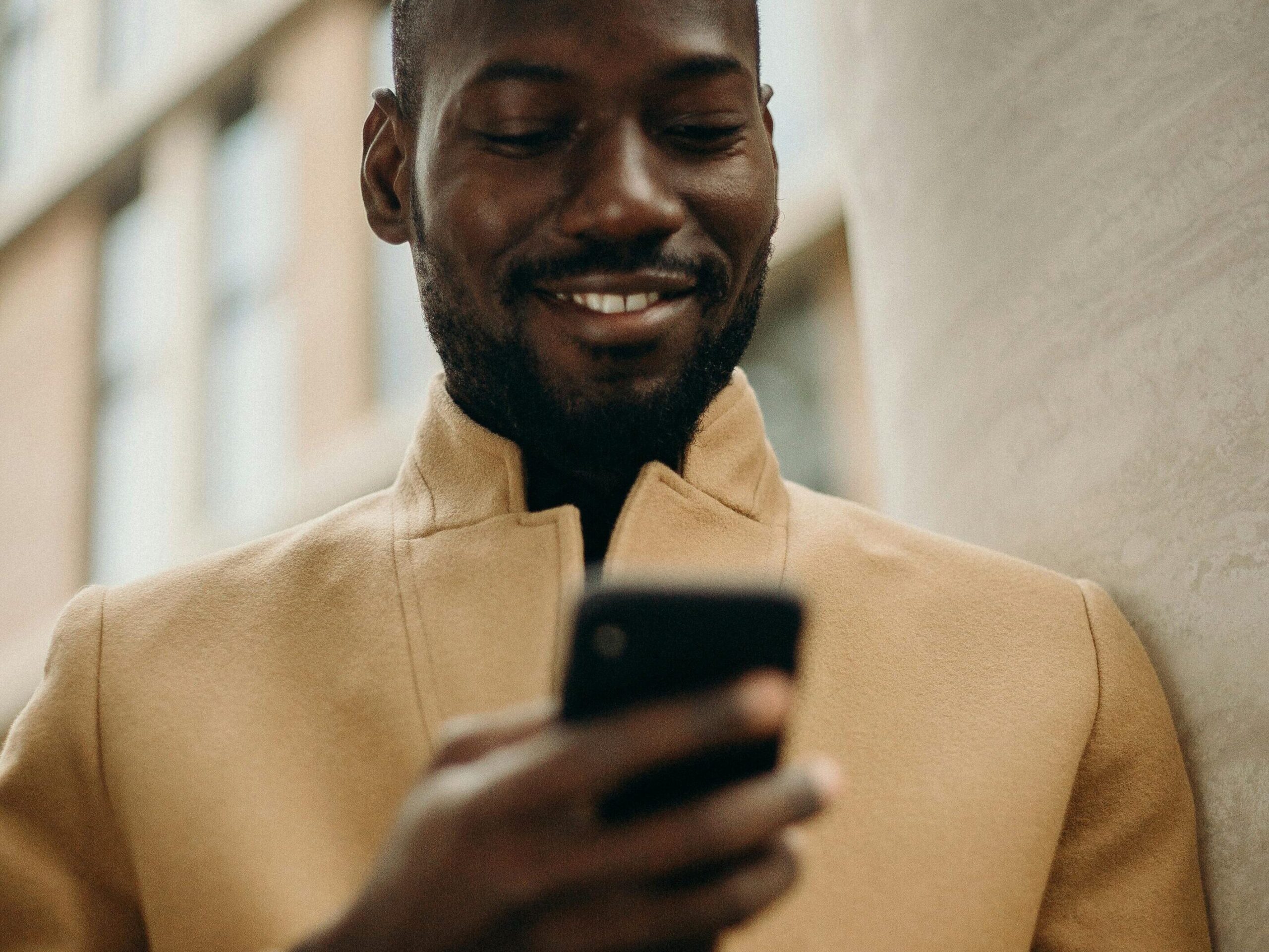 A cheerful man in a stylish coat using his smartphone and holding coffee outside.
