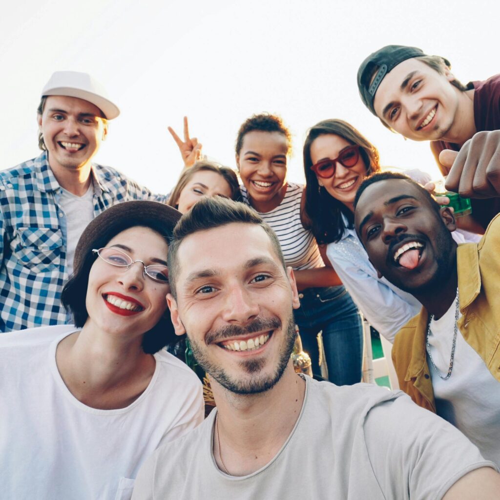 A diverse group of friends smiles while taking a selfie outdoors during the day.